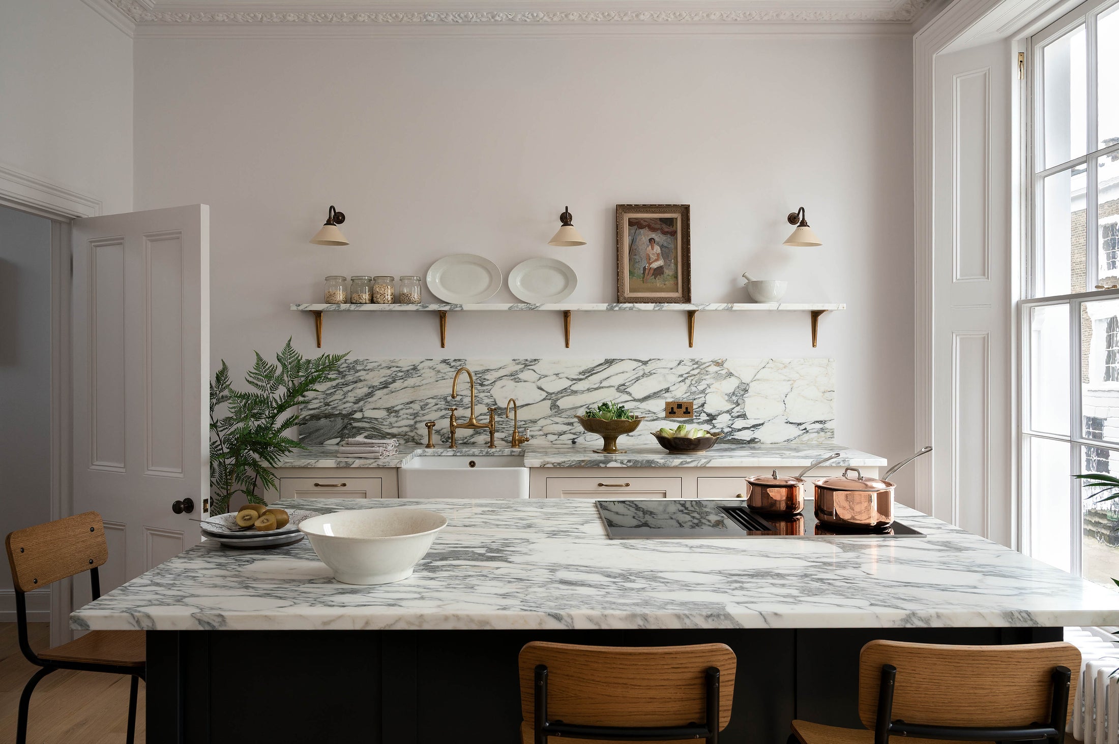 A humble yet elegant off-white Classic English kitchen features a sink run with honed Arabescato marble worktop and splashback, a pantry blue kitchen island with a cooker, mini creamware wall lights, and plumber’s stools. Adorned with artisan ceramics, brass pans, a vintage painting, fresh greens, a