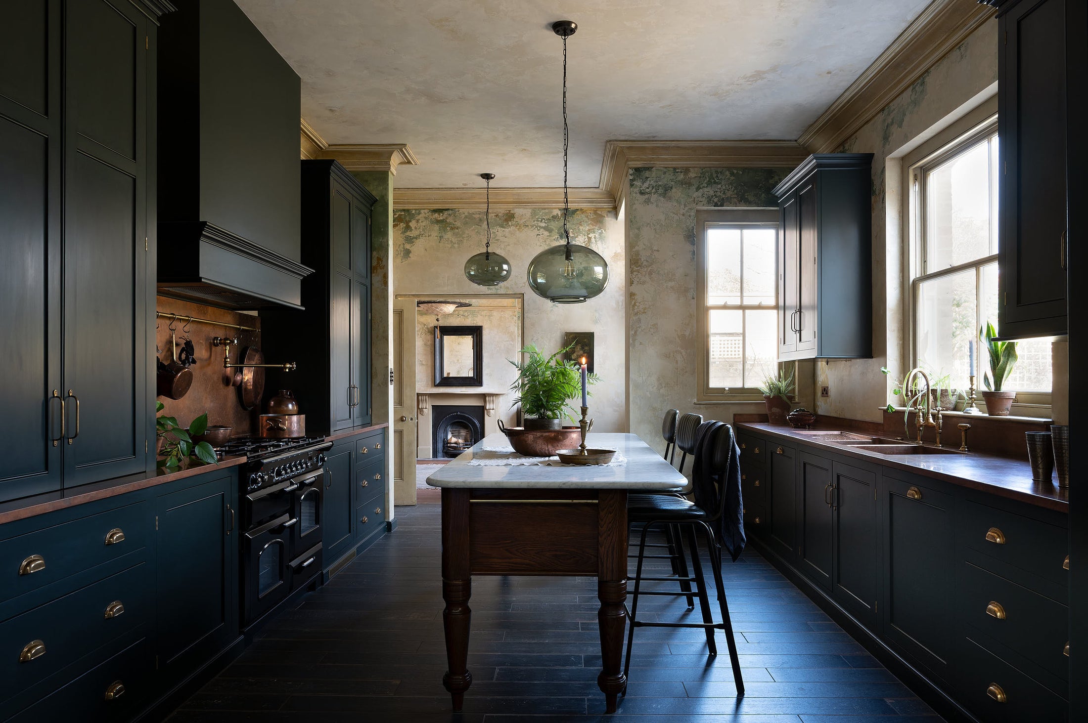 An elegant classic kitchen includes a cooker and sink with an aged copper worktop, a shaker canopy, counter cabinets, a marble worktop heirloom dairy table, a breakfast nook, and tall black chairs. The tall-ceilinged room has a rustic wash finish, with green draping down the walls, resembling overgr