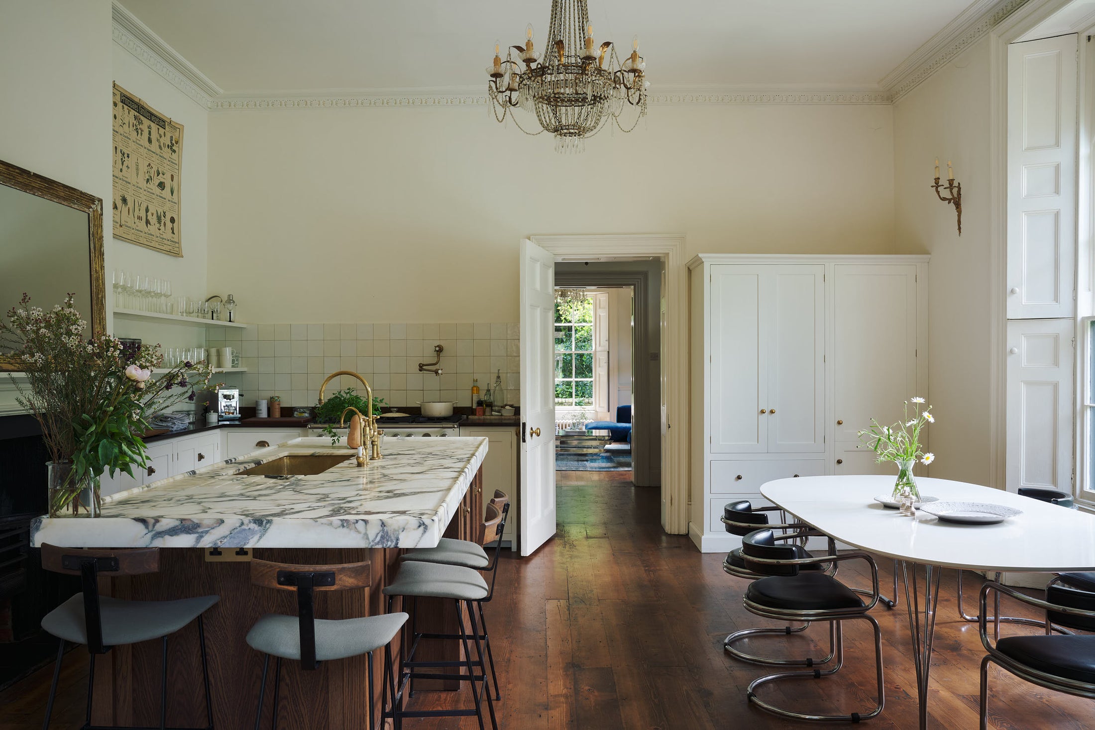 A classic English kitchen in white linen features white and cream splashback tiles, a cooker with a polished sapelle worktop, and a kitchen island topped with Arabescato marble. It includes a polished brass sink, an aged brass Ionian tap, a scientific illustration of vegetable species, two glasses o