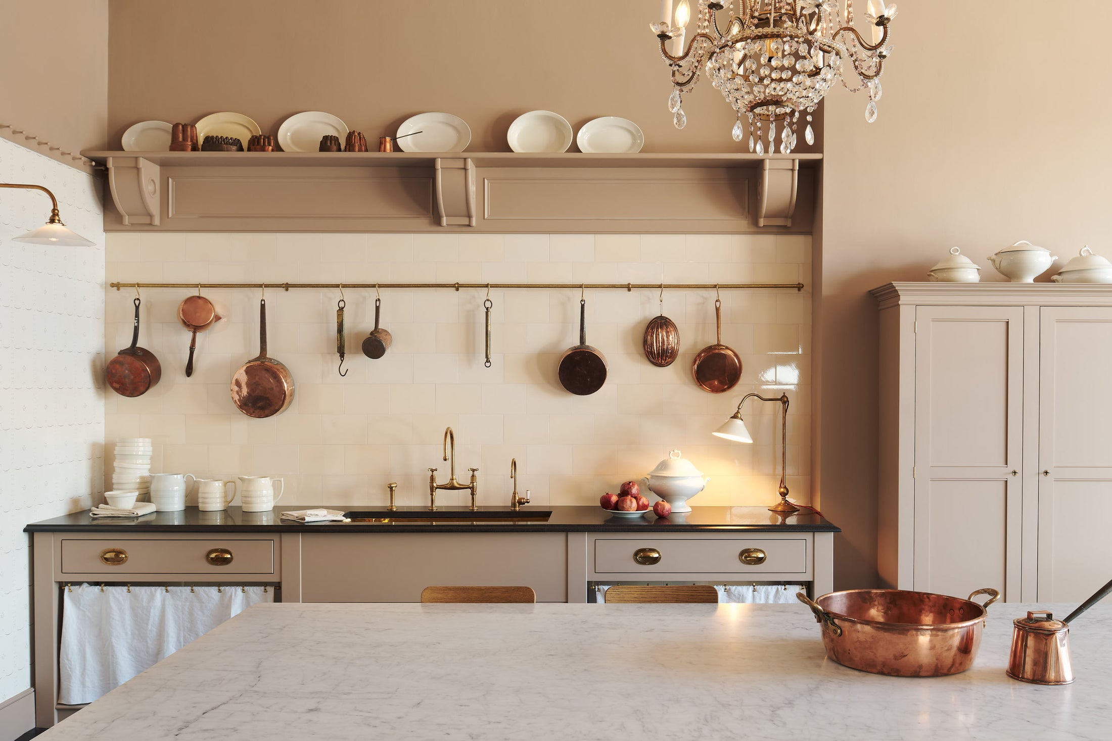 A grand Princelet pink Heirloom and Classic English kitchen features a sink cabinet with a black granite worktop, a butler’s pantry, a large kitchen island with a Carrara marble worktop, aged brass rails, ditsy delft wallpaper, a tall cream subway tile splashback, a grand tour desk lamp, and an Heir