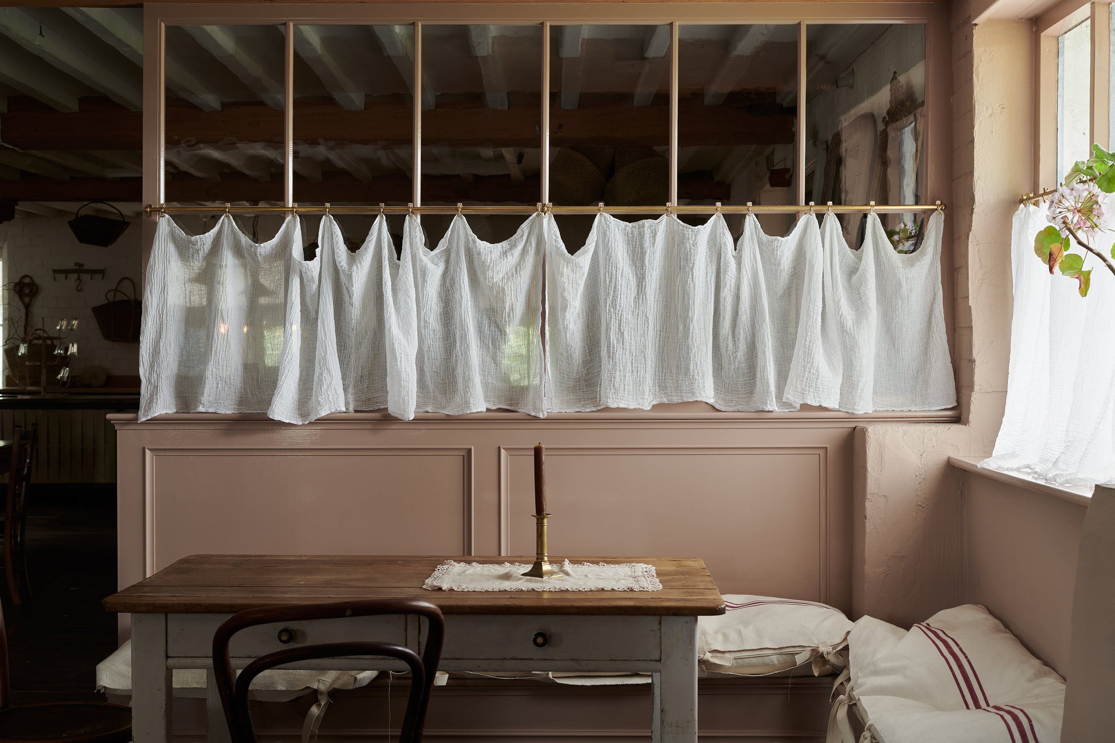 Soft muslin fabric is gently draped from small brass clips on an Aged Brass Cafe Curtain Rail in front of a half-glazed partition wall painted in a dusty pink hue. In the foreground is an antique table, a glimpse of a Nacoco Cafe Chair, and a single brass candle stick, creating a cosy seating area.