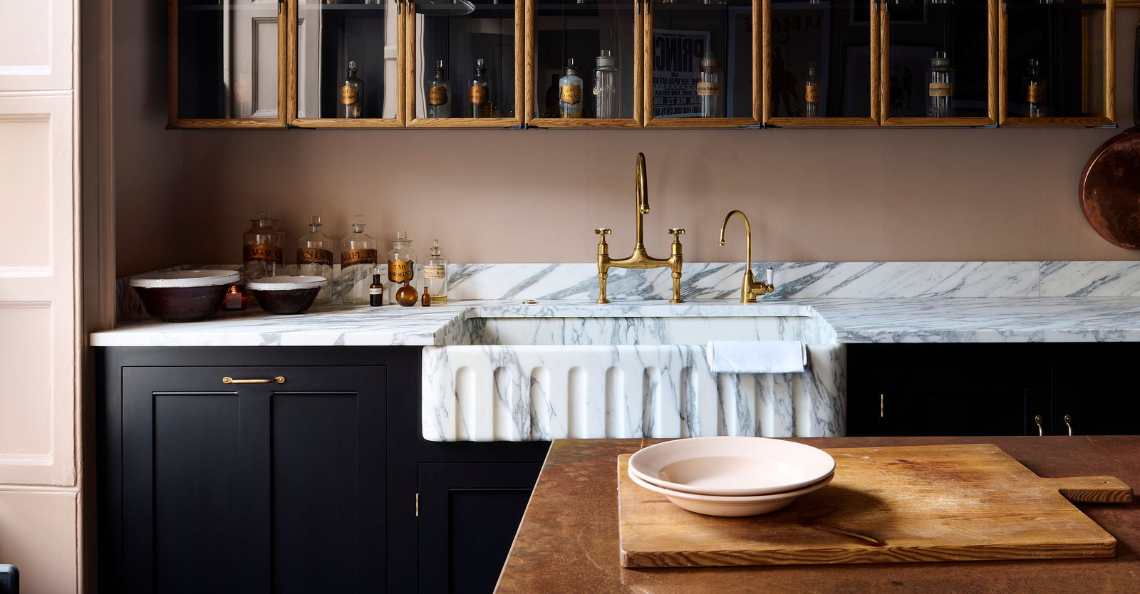 A bold and dramatic Haberdasher’s kitchen with a sink and cooker run, an Arabescato worktop and single fluted sink, an aged brass Ionian tap, a glazed wall cupboard, a large copper canopy, and a kitchen island with a copper worktop. Adorned with copper pans, artisan ceramics, labelled glass jars, an