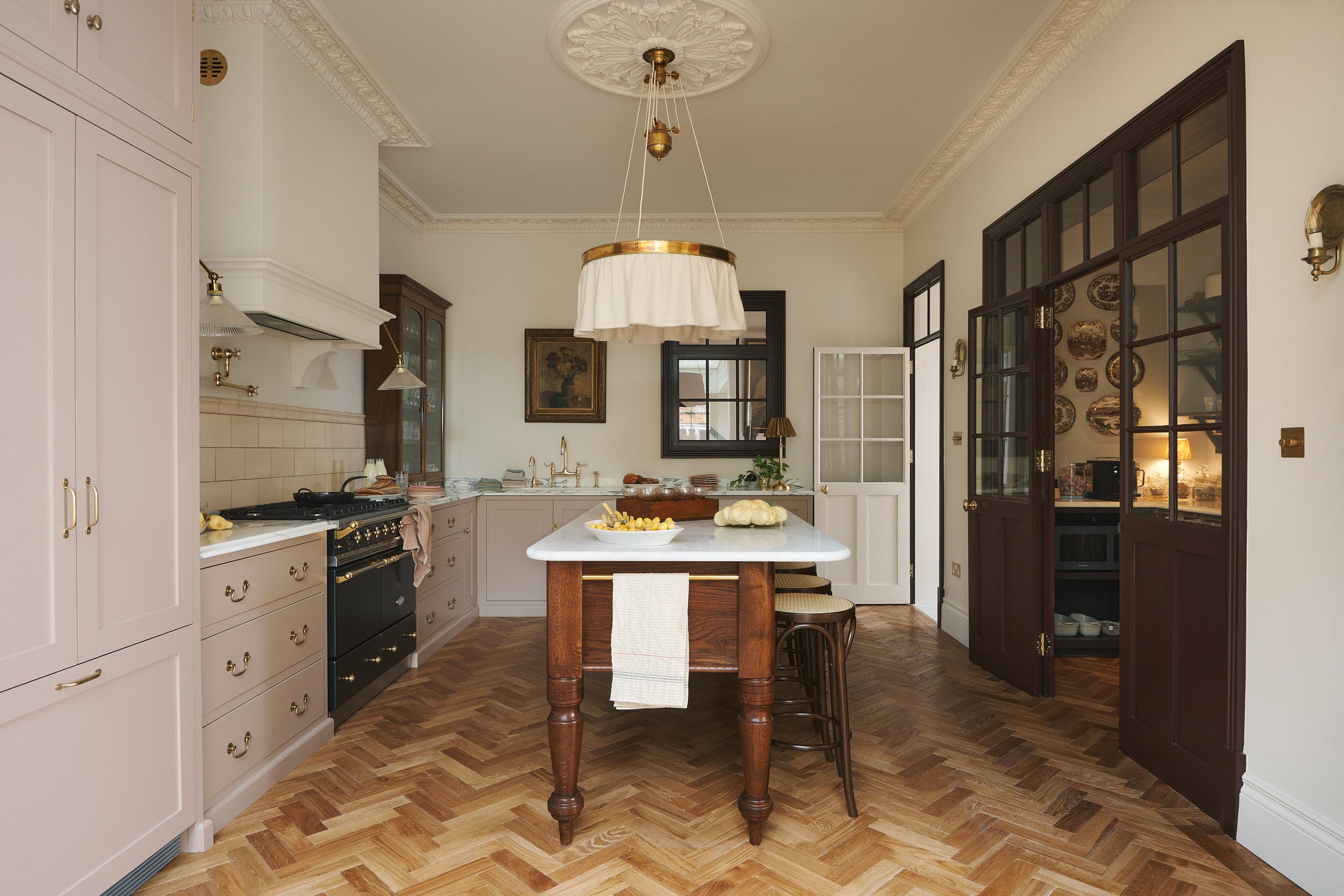 A soft Princelet pink Shaker kitchen unfolds with an L-shaped cooker and sink run topped in Carrara marble. It features a counter cupboard, full-height cabinets, cream subway tiles, a large Dairy Table surrounded by four Bentwood café stools, and glazed doors leading to a spacious pantry. Gold accen