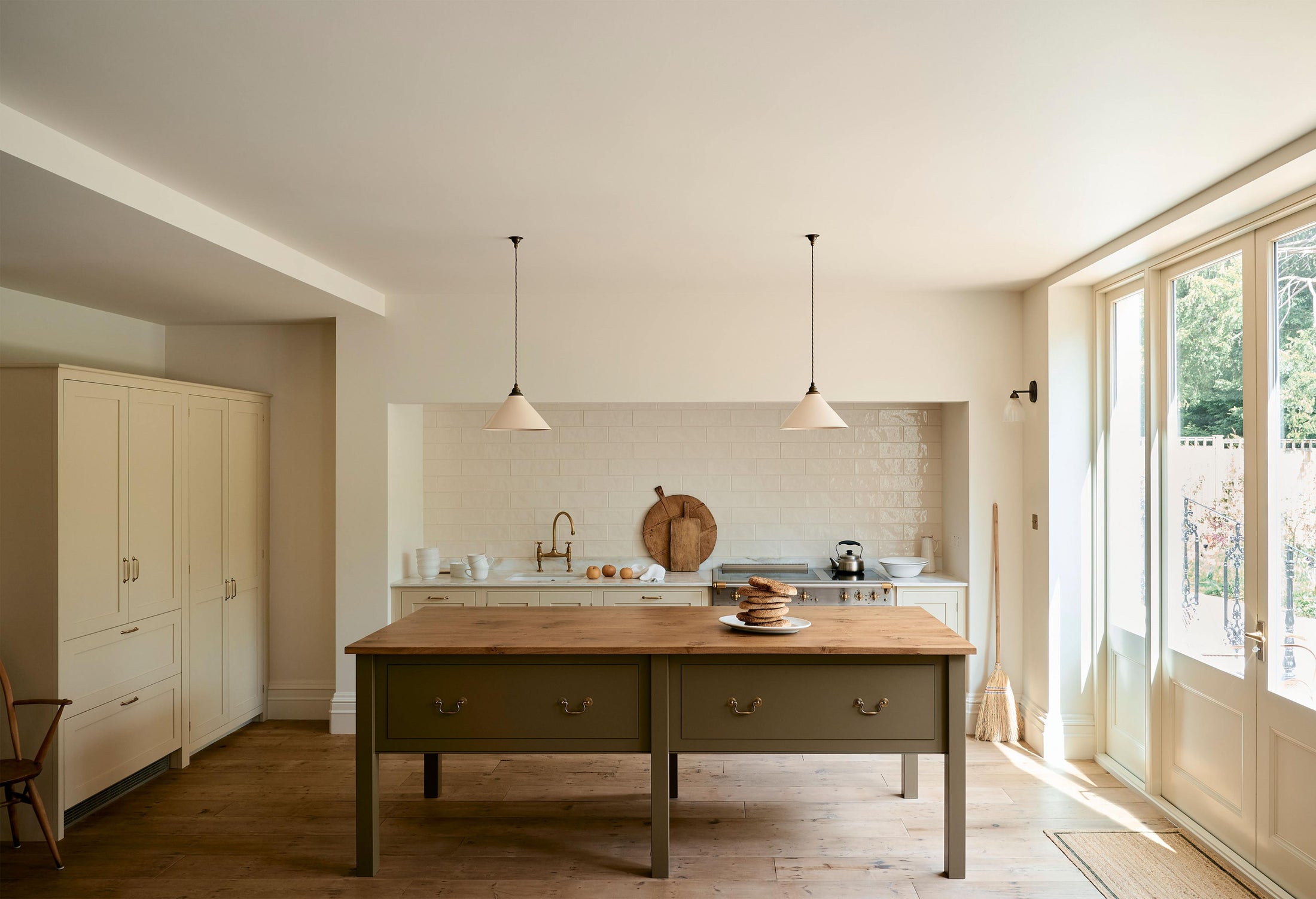 A light, clean, and simple shaker kitchen features a sink and cooker run nestled in a chimney breast, cream porcelain splashback tiles, wooden floorboards, and white walls. Cream cabinets complement an olive green Georgian-style prep table with deep drawers and a rustic oak worktop at the room’s cen